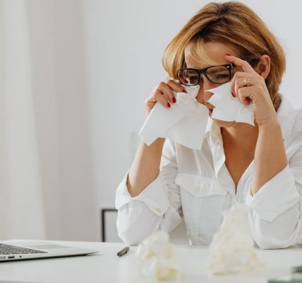 A woman in glasses is wiping tears while seated at a desk with a laptop and tissues.