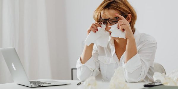 A woman in glasses is wiping tears while seated at a desk with a laptop and tissues.